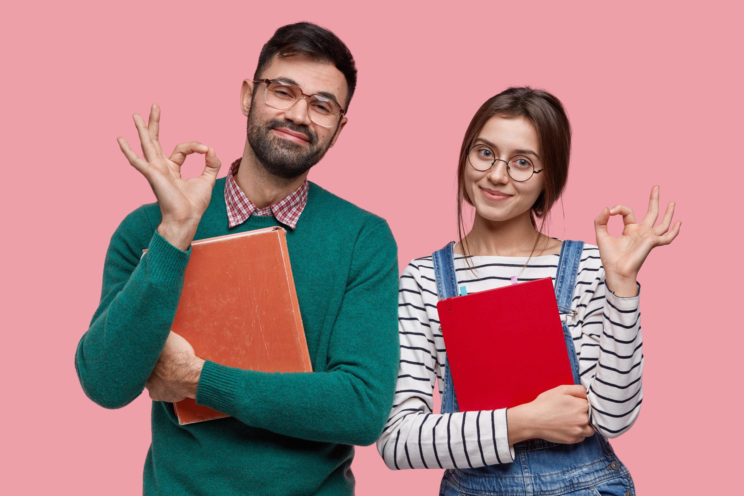 Photo of handsome male student and his female groupmate demonstrates okay gesture, agree with something, hold book and notebook closely, isolated over pink background
